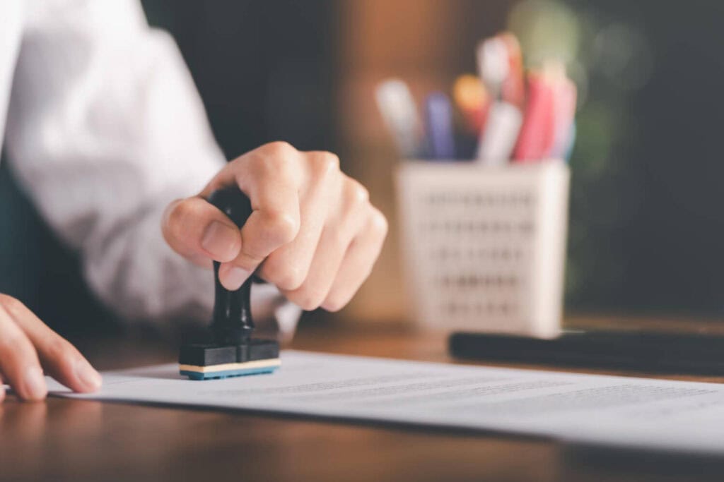 Nahaufnahme einer Hand, die einen schwarzen Stempel auf ein Blatt Papier drückt. Die Person trägt ein weißes Hemd. Das Papier liegt auf einem Holztisch und enthält gedruckten Text in kleiner Schrift. Im Hintergrund ist ein unscharfer Behälter mit mehreren farbigen Stiften sowie ein dunkler länglicher Gegenstand (vermutlich ein Stift) zu sehen. Die Szene ist in warmem Licht gehalten.