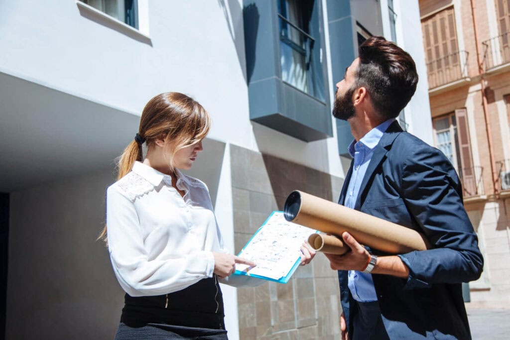 Auf dem Bild sieht man zwei Personen, die eine Immobilienbewertung oder Besichtigung durchführen. Die Frau hält einen Klemmbrett mit Plänen oder Unterlagen in der Hand und zeigt auf etwas, während der Mann mit Bauplänen im Arm die Fassade des Gebäudes betrachtet.